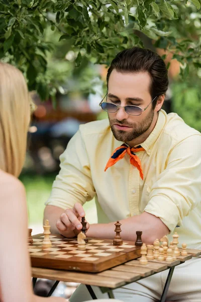 Hombre con estilo en gafas de sol jugando ajedrez con mujer borrosa en el parque de verano - foto de stock