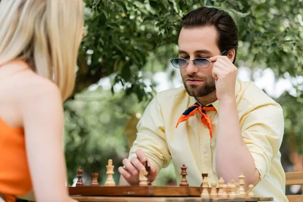 Young man holding sunglasses near chess and blurred girlfriend in park — Stock Photo