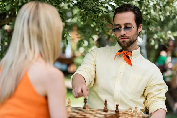 Trendy young man pointing at chess figure near blurred girlfriend in summer park — Stock Photo