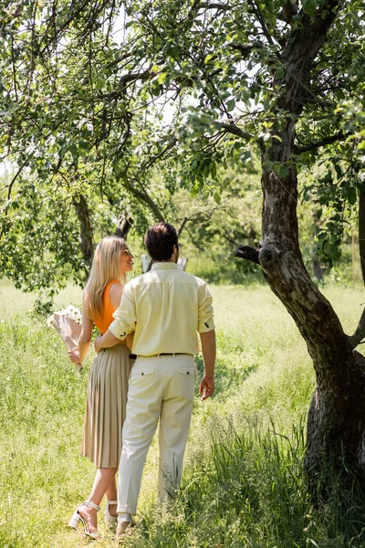 Donna sorridente in occhiali da sole che tiene bouquet mentre cammina con il fidanzato nel parco estivo — Foto stock