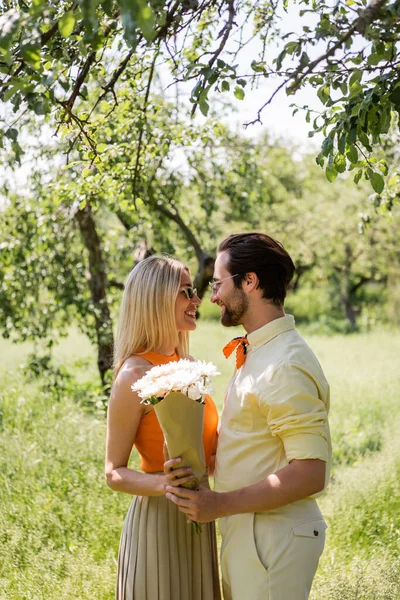 Vista lateral do casal elegante em óculos de sol segurando flores no parque de verão — Fotografia de Stock