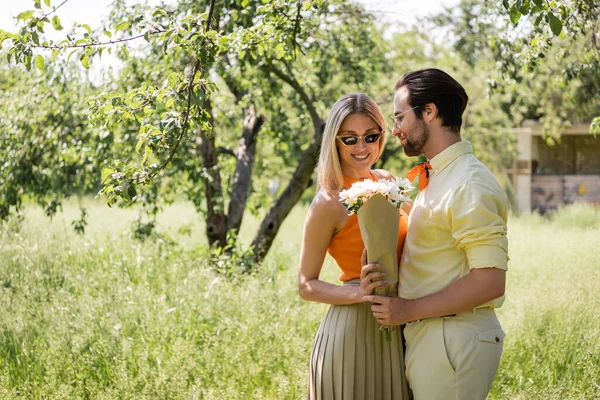 Homme à la mode dans les lunettes de soleil tenant bouquet près de petite amie souriante dans le parc d'été — Photo de stock