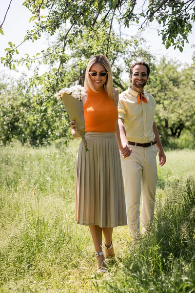 Mujer alegre y elegante en gafas de sol sosteniendo flores mientras camina con su novio en el parque de verano - foto de stock