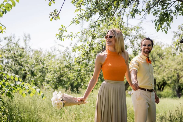 Mujer feliz sosteniendo ramo y mano de novio de moda en el parque de verano - foto de stock