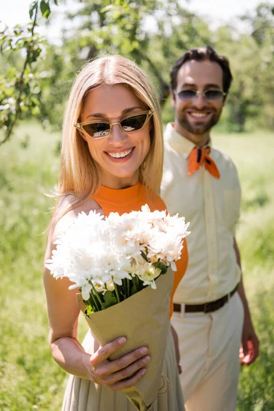 Mujer de moda mirando crisantemos cerca de novio borroso en el parque - foto de stock