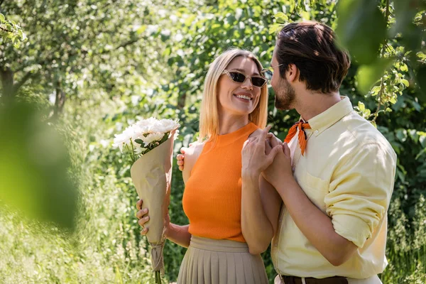 Cheerful woman holding chrysanthemums and hand of young boyfriend in summer park — Stock Photo