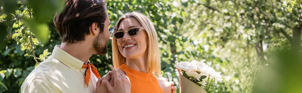 Позитивная женщина держит букет рядом с парнем в солнцезащитных очках в летнем парке, баннер — стоковое фото