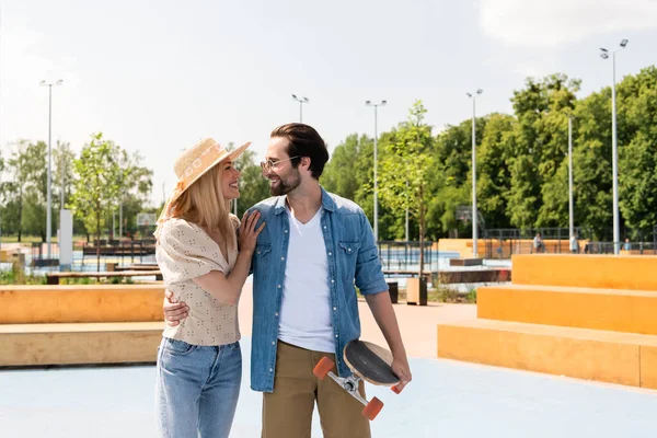 Vista lateral da mulher sorridente abraçando o jovem namorado com longboard no parque de skate — Fotografia de Stock