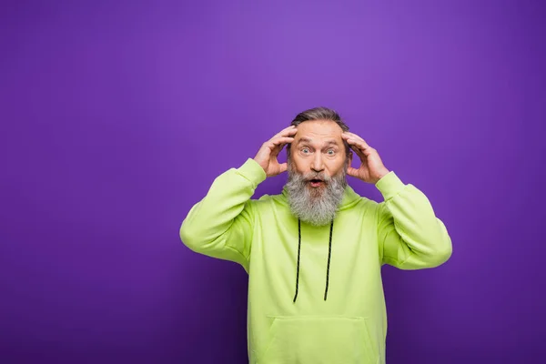 Shocked and bearded senior man with grey hair looking at camera on purple — Stock Photo