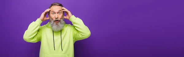 Shocked and bearded senior man with grey hair looking at camera on purple, banner — Stock Photo