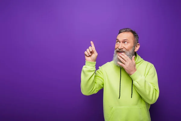 Hombre mayor feliz y barbudo con el pelo gris mirando hacia otro lado mientras señala con el dedo en púrpura - foto de stock