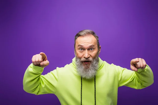 Überraschte Seniorin mit Bart und grauen Haaren zeigt mit Fingern auf lila Kamera — Stockfoto