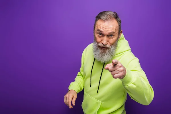 Senior Mann mit Bart und grauen Haaren zeigt mit dem Finger auf Kamera auf lila — Stockfoto