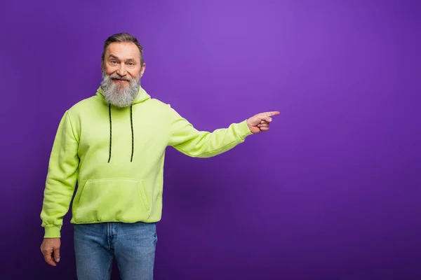 Homem sênior feliz com barba e cabelos grisalhos em pé com capuz verde e apontando com o dedo no roxo — Fotografia de Stock