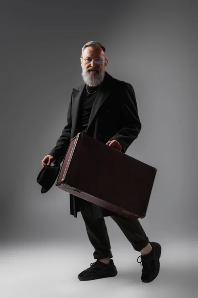 Full length of elegant senior man in black coat holding derby hat and suitcase on grey — Stock Photo