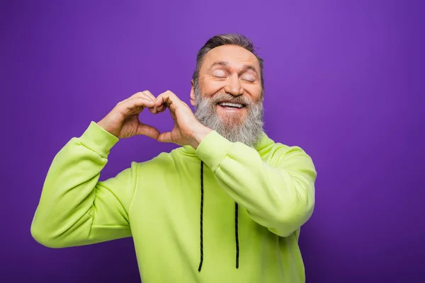 Cheerful senior man with closed eyes and grey hair showing heart with hands on purple — Stock Photo