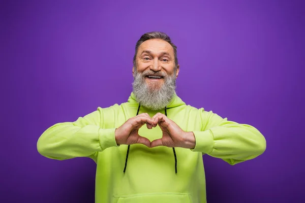 Fröhlicher älterer Mann mit Bart und grauen Haaren, der Herz mit Händen auf lila zeigt — Stockfoto