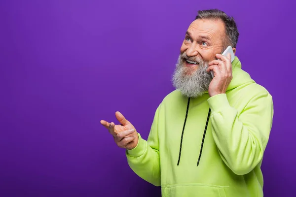 Glücklicher älterer Mann mit Bart und grauen Haaren, der auf dem Smartphone auf lila spricht — Stockfoto