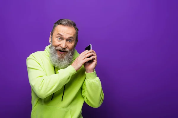 Verblüffter Senior mit Bart und grauen Haaren hält Smartphone auf lila — Stockfoto