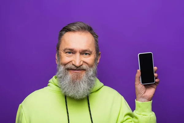 Heureux homme âgé avec barbe et cheveux gris tenant smartphone avec écran vide isolé sur violet — Photo de stock