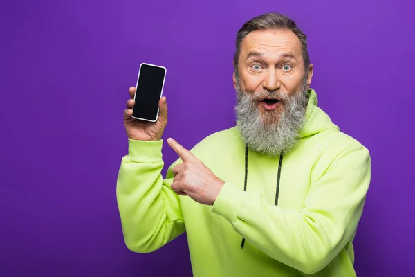 Amazed senior man with beard and grey hair pointing at smartphone isolated on purple — Stock Photo