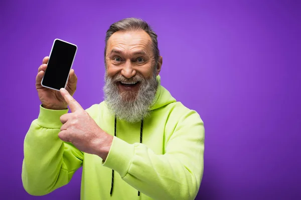 Happy senior man with beard and grey hair pointing at smartphone on purple — Stock Photo