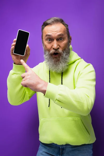 Sorprendido hombre mayor con barba y pelo gris apuntando a teléfono inteligente en púrpura - foto de stock