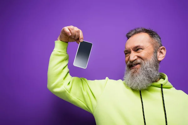 Unzufriedener älterer Mann mit Bart und grauen Haaren hält Smartphone mit leerem Bildschirm auf lila — Stockfoto