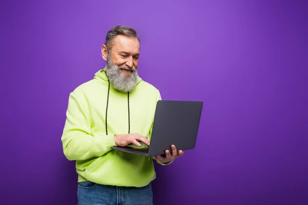 Heureux homme âgé avec barbe et cheveux gris en utilisant un ordinateur portable sur violet — Photo de stock