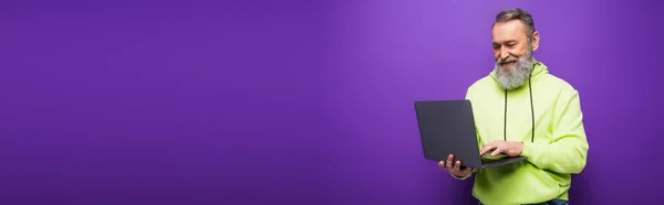 Happy senior man with beard and grey hair using laptop on purple background, banner — Stock Photo