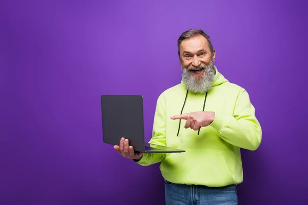 Heureux homme âgé avec barbe et cheveux gris pointant avec le doigt à l'ordinateur portable sur violet — Photo de stock