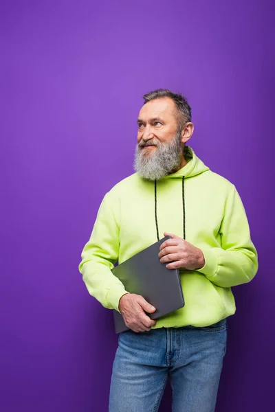 Happy senior man with beard and grey hair holding laptop while standing and looking away on purple — Stock Photo