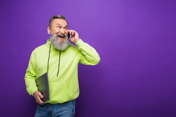 Happy senior man with beard and grey hair holding laptop while talking on mobile phone on purple — Stock Photo