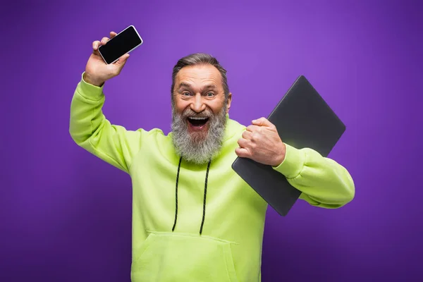 Hombre mayor feliz con barba y pelo gris celebración portátil y teléfono inteligente con pantalla en blanco en púrpura - foto de stock