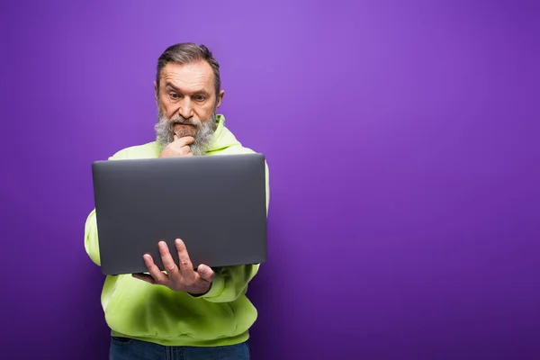 Homem sênior pensativo com barba e cabelos grisalhos usando laptop no fundo roxo — Fotografia de Stock