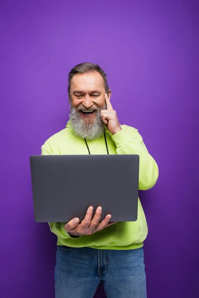 Positiver älterer Mann mit Bart und grauen Haaren mit Laptop auf lila — Stockfoto