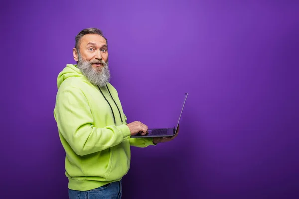 Sorridente homem sênior com barba e cabelos grisalhos usando laptop no fundo roxo — Fotografia de Stock