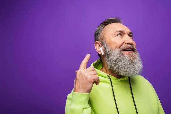 Felice uomo anziano con cappuccio verde ascoltare musica e puntare con il dito al auricolare wireless viola — Foto stock