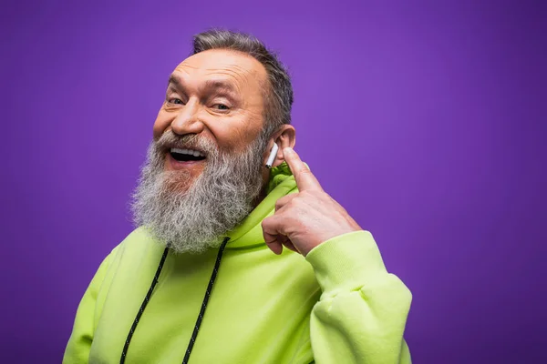 Cheerful senior man in green hoodie listening music and pointing with finger at wireless earphone on purple — Stock Photo