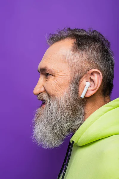 Side view of happy senior man in green hoodie listening music in wireless earphone on purple — Stock Photo