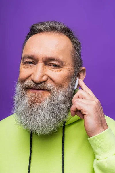 Happy senior man in green hoodie listening music and adjusting wireless earphone on purple — Stock Photo