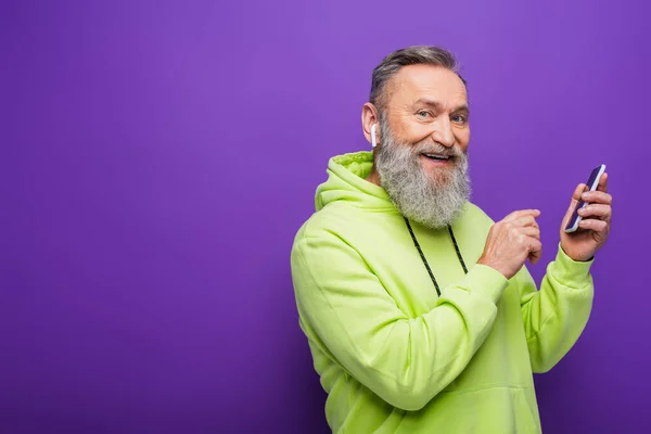 Happy senior man in green hoodie listening music in wireless earphone and holding smartphone on purple — Stock Photo