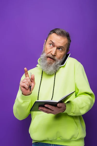 Senior man in green hoodie holding pen and notebook while talking on smartphone on purple — Stock Photo
