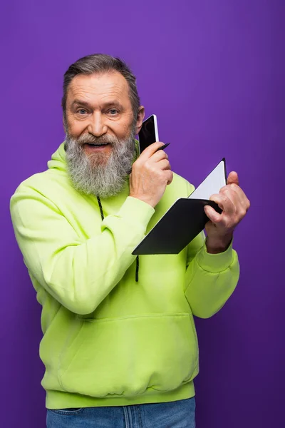 Happy senior man in green hoodie holding notebook and talking on smartphone on purple — Stock Photo
