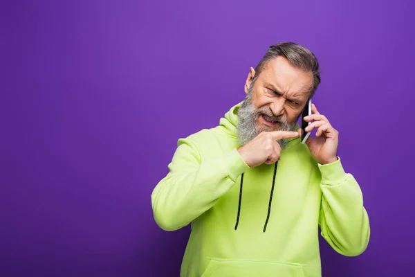 Angry senior man in green hoodie pointing with finger while talking on smartphone on purple — Stock Photo