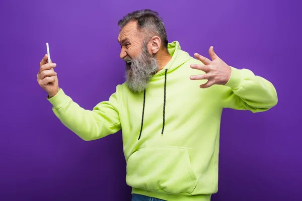 Stressed senior man in green hoodie looking at smartphone on purple — Stock Photo