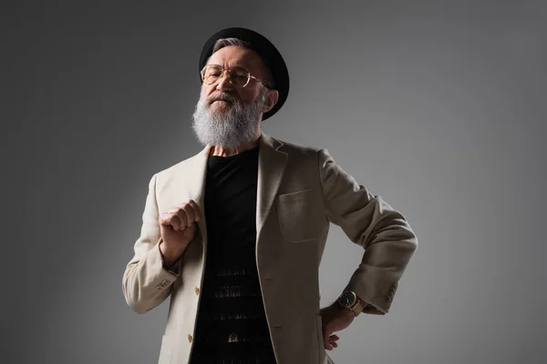 Stylish senior man in beige jacket and derby hat posing with hand on hip on grey — Stock Photo