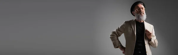 Stylish senior man in beige jacket and derby hat posing with hand on hip on grey, banner — Stock Photo