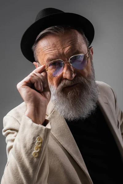 Stylish senior man in beige jacket and derby hat adjusting eyeglasses on grey — Stock Photo