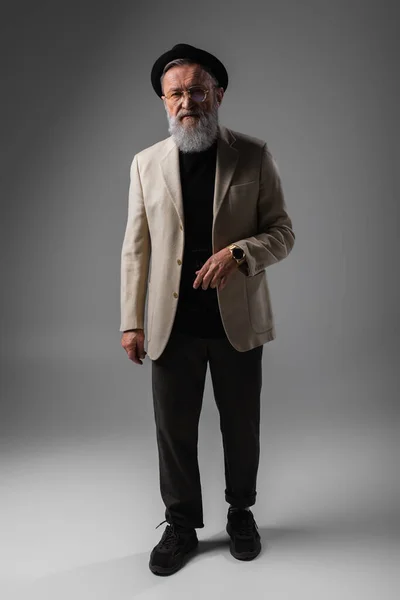 Full length of stylish senior man in beige jacket and derby hat standing on grey — Stock Photo